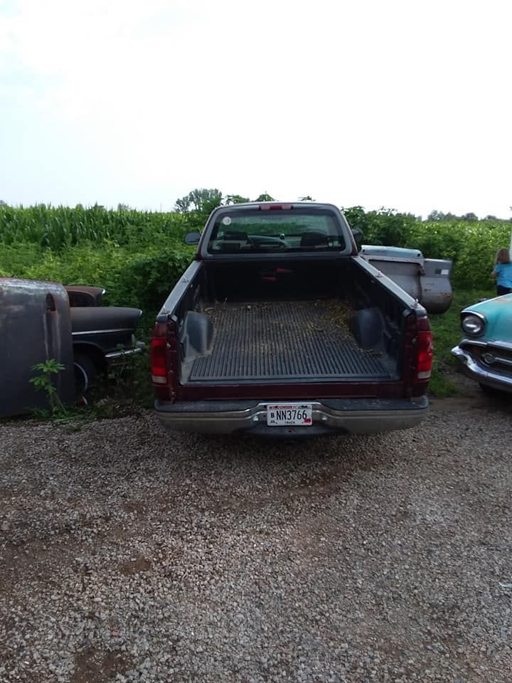 Rear view of a maroon pickup truck with an open bed, parked on gravel with old cars nearby.