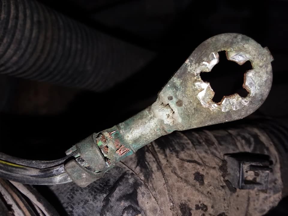 Corroded electrical wire connector with a star-shaped hole, attached to a black cable in a vehicle's engine.