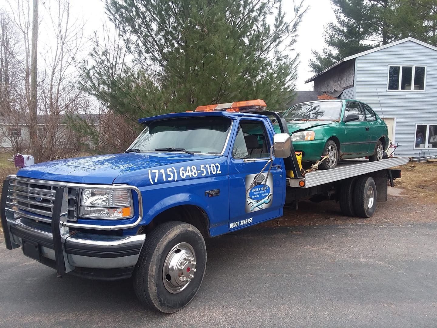 Blue tow truck hauling a green car on a gray, overcast day.  The truck has a front bumper guard.