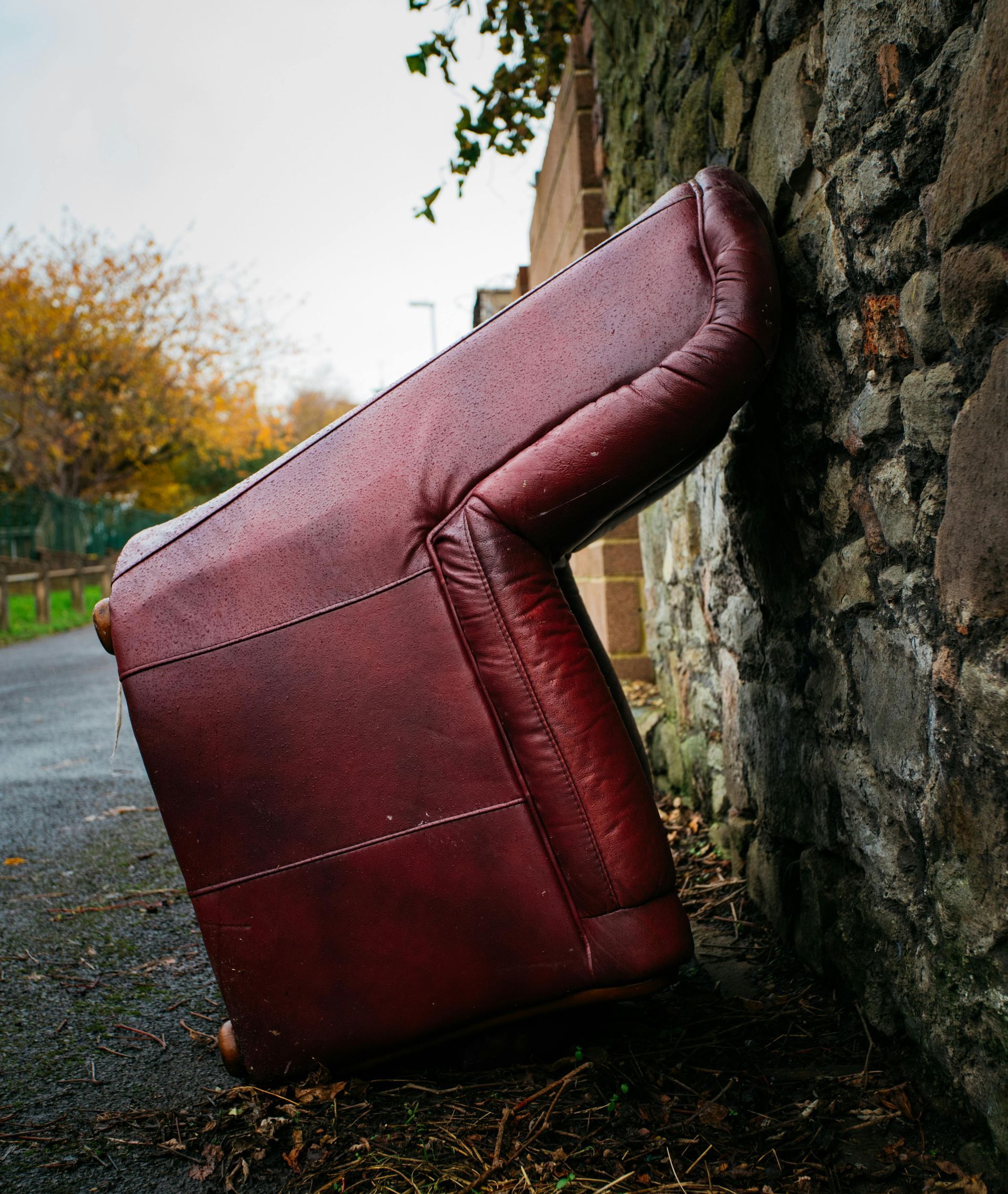 A maroon leather armchair leaning against a stone wall on a wet sidewalk.