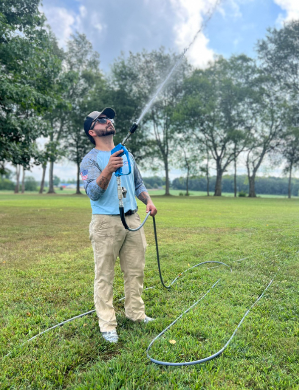 Person wearing a mask sprays water on grass in a park, holding a hose under a cloudy sky.