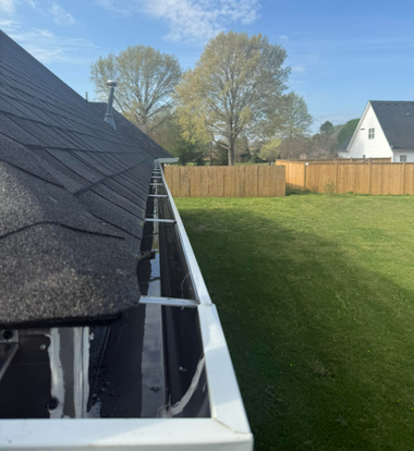 Residential backyard with a roof edge and gutter in the foreground, lawn, fence, and trees under a blue sky