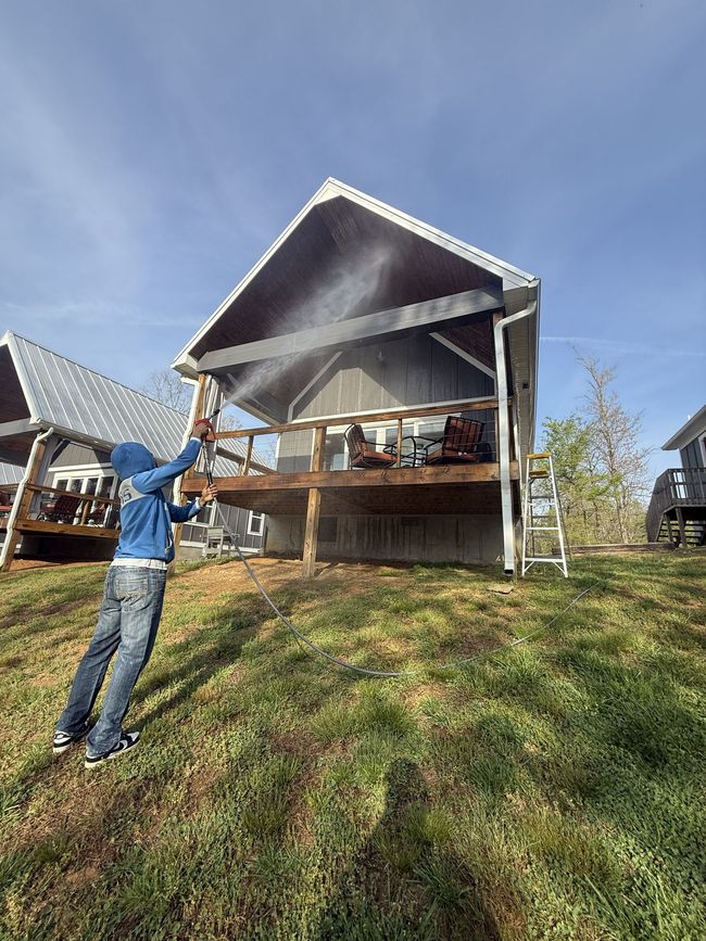 Person spraying water on a smoking cabin porch on a grassy hill under a blue sky