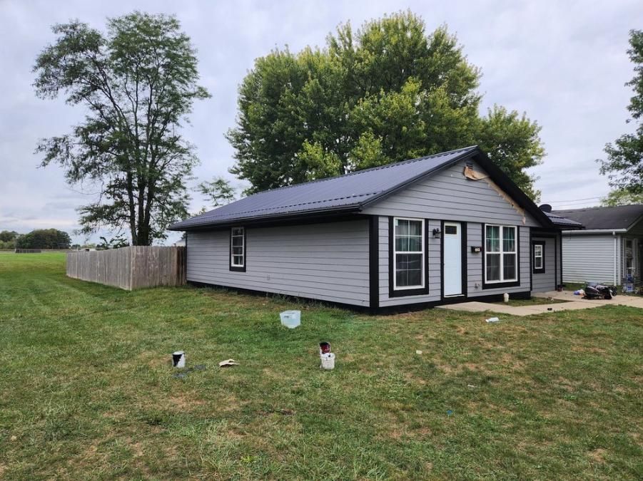 Small gray house with black roof in a grassy yard under a cloudy sky
