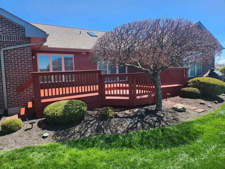 Brick house with red wooden deck, trimmed shrubs, and a small tree in a sunny yard.
