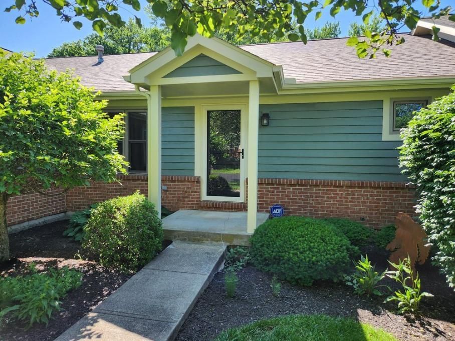 Front entry of a small green house with brick base, white trim, and a concrete walkway through landscaped shrubs.
