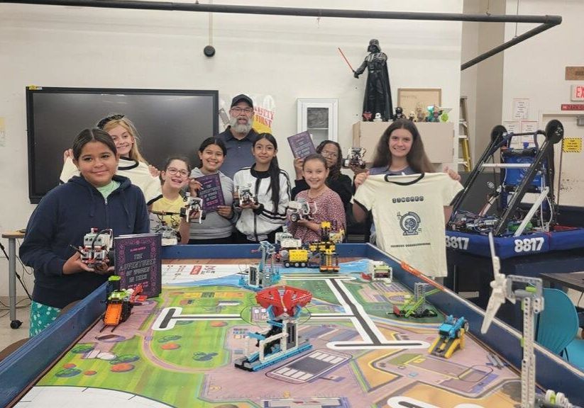 Group of kids posing behind a robotics competition table with a small robot and game field indoors