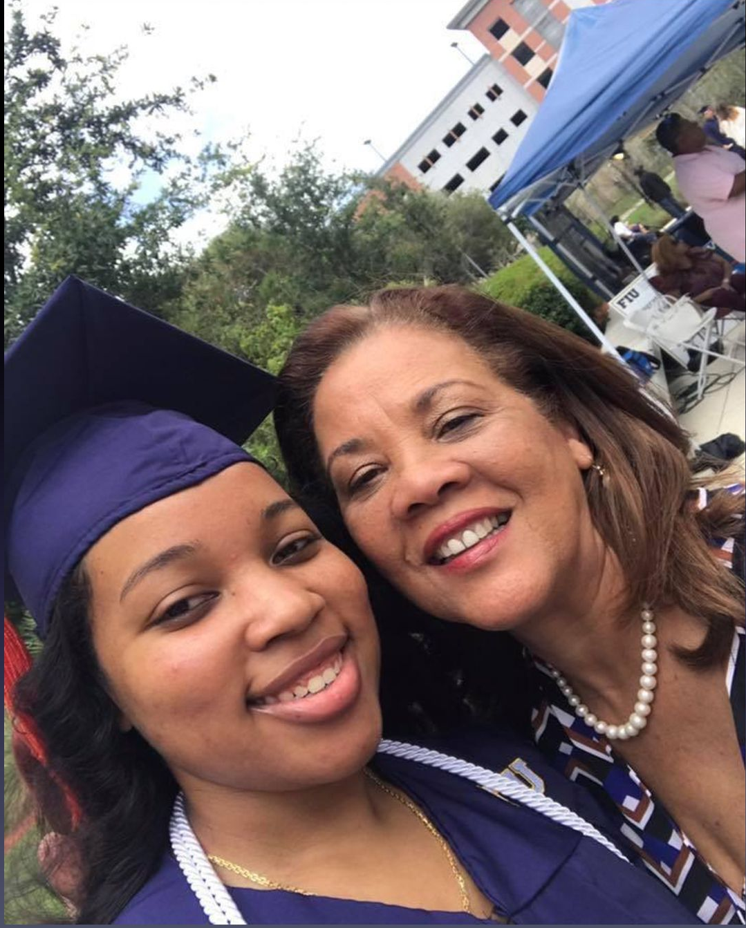 Two people smiling at an outdoor event, one in a graduation cap and gown.