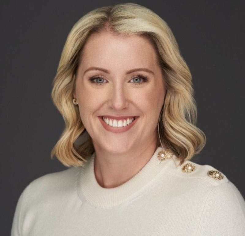 Smiling blonde woman in a white sweater with gold buttons against a dark background
