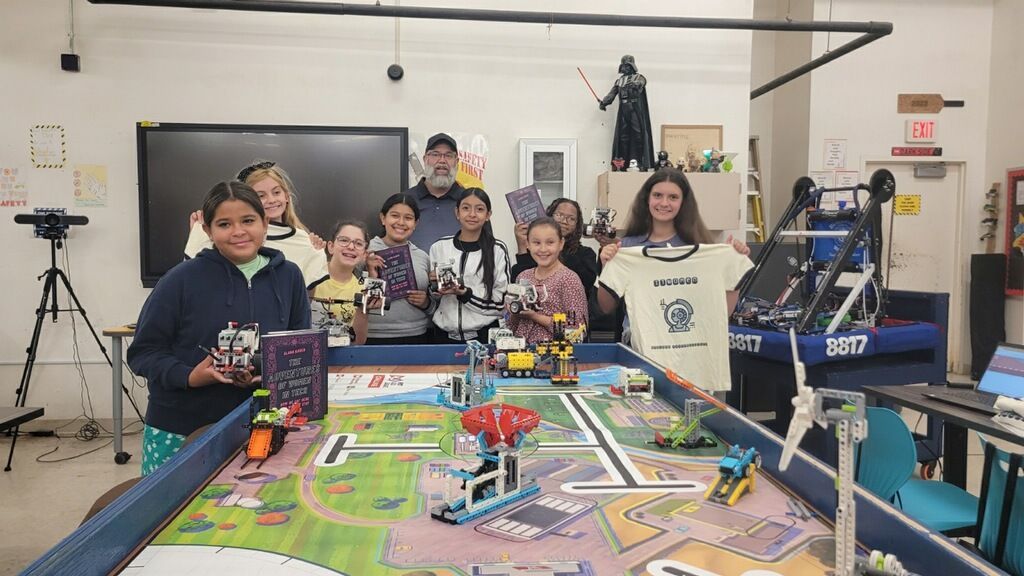 Team posing behind a colorful LEGO robotics table in a workshop room, smiling with trophies and equipment.