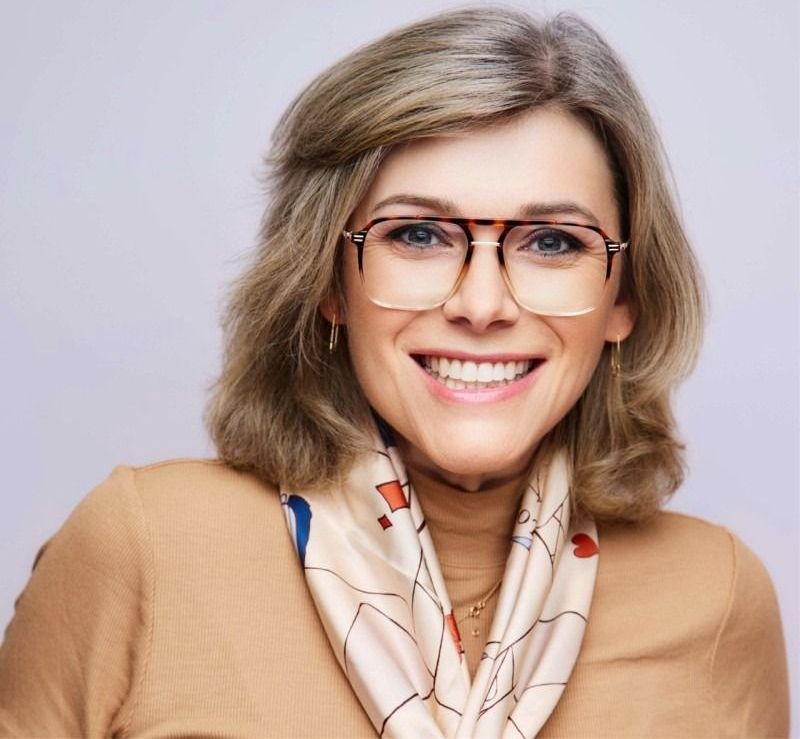 Smiling woman wearing glasses and a beige scarf against a light background