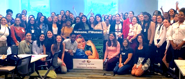 Large group posed indoors with a banner, smiling and seated around a central plaque or sign.