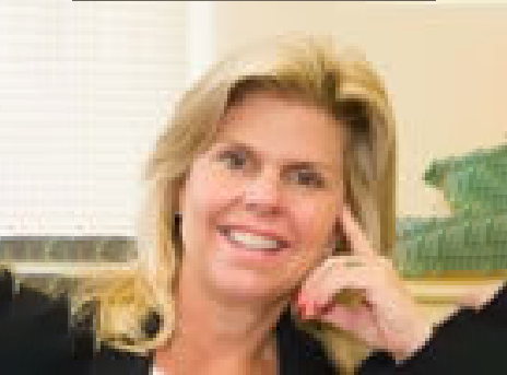 Smiling blonde woman seated indoors, resting her head on her hand.