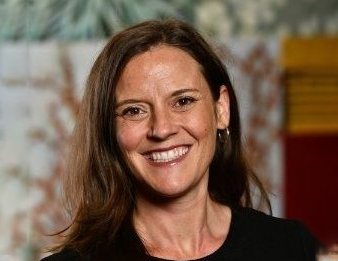 Smiling woman with shoulder-length brown hair in a black top against a blurred indoor background