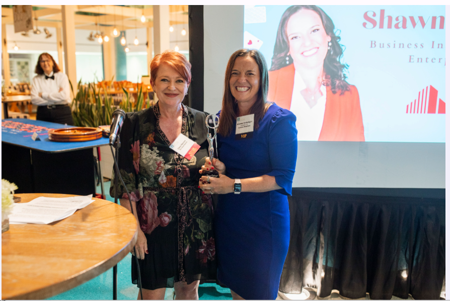 Two women pose at a conference table with a projected presentation in the background.