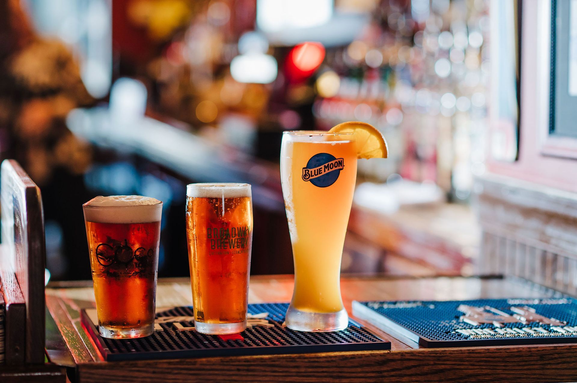 Three iced drinks on a bar tray, including a tall orange cocktail with a fruit garnish.