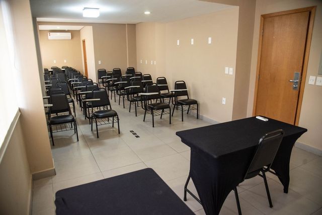 Sala de reuniões vazia com fileiras de cadeiras pretas e duas mesas com toalhas pretas sob luzes quentes.