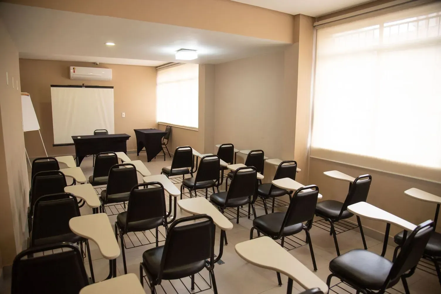 Sala de aula vazia com fileiras de cadeiras e mesas, uma tela de projeção e janelas bem iluminadas ao longo da parede direita.