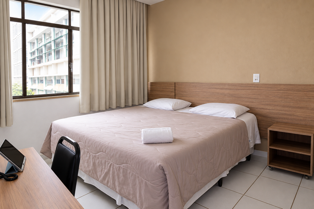 Quarto pequeno com cama, escrivaninha, janela e cortinas bege, visto da porta.