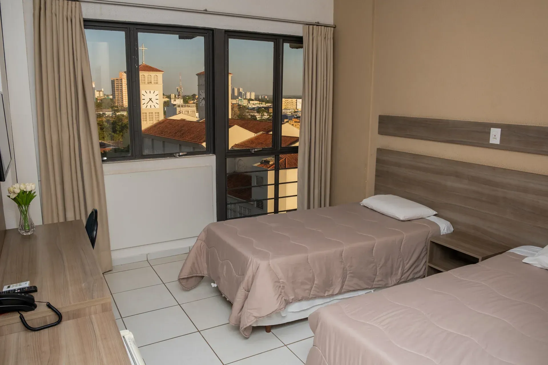 Quarto com duas camas de solteiro, escrivaninha e portas de varanda com vista para os telhados.
