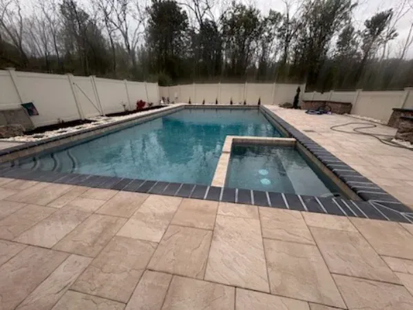 Outdoor backyard pool with attached hot tub, surrounded by beige stone patio and white privacy walls