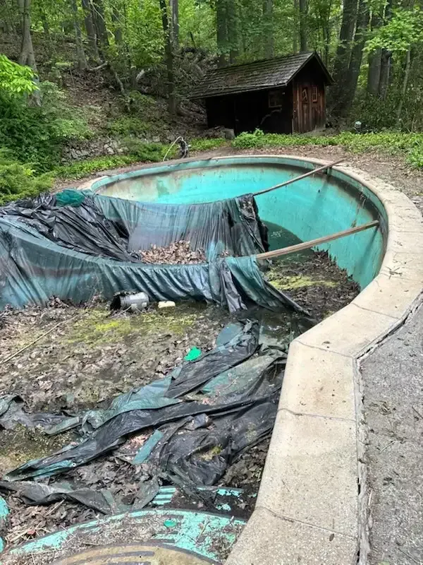 Partially drained backyard pool with torn black tarp and turquoise liner, surrounded by woods and a concrete edge