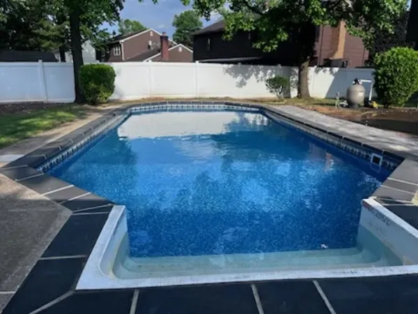 Backyard swimming pool with blue water, concrete deck, and a white privacy fence under trees