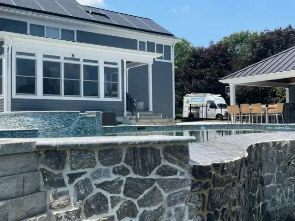 Stone patio with a pool, gray house, and covered outdoor dining area under a sunny sky