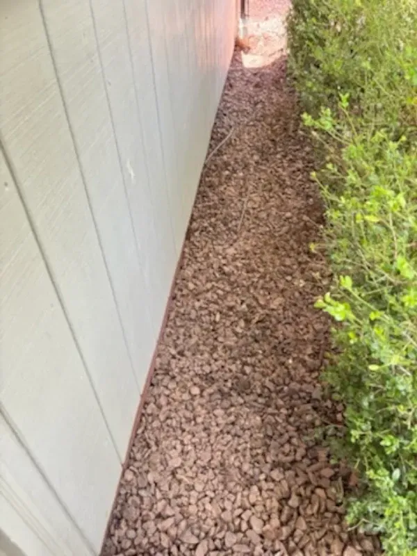 Narrow gravel strip beside a white wall and green bushes along a building exterior