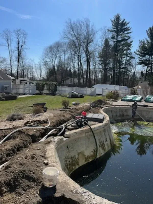 Backyard pond under construction with exposed dirt, hoses, and trees in the background