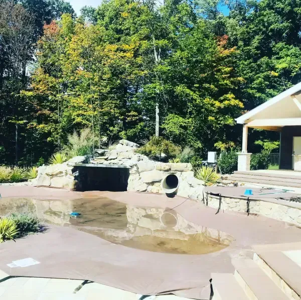 An empty outdoor pool with muddy water, a rock waterfall, and a pavilion beside trees.