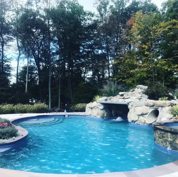 Backyard swimming pool with blue water, stone grotto slide, and trees surrounding it