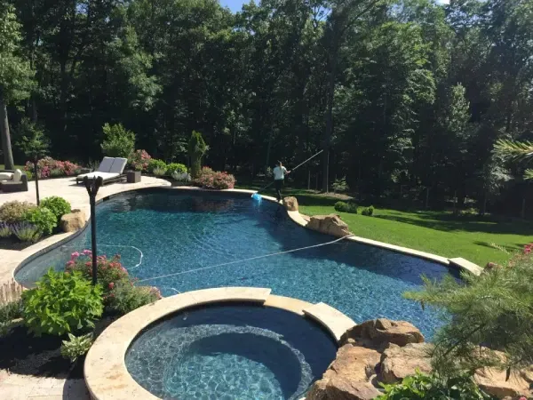 Backyard pool with a curved spa, blue water, rocks, and surrounding trees and lawn