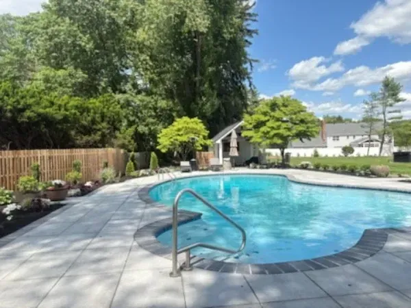 Backyard pool with blue water, metal handrail, stone deck, trees, and a small pool house on a sunny day