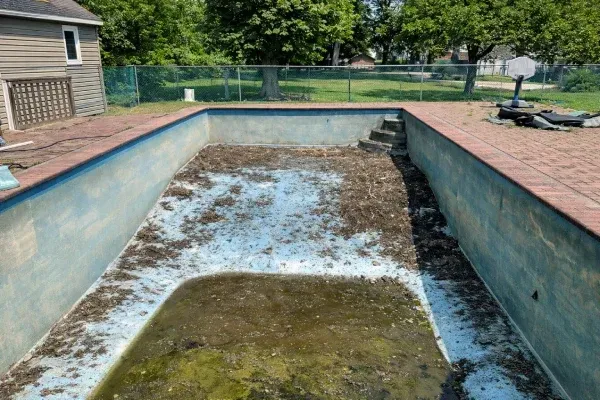 Empty, weathered backyard swimming pool with standing water and debris, surrounded by brick coping and a fence