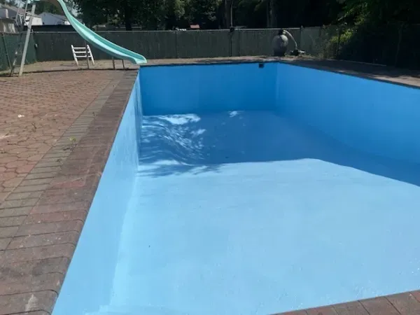 Empty blue swimming pool beside a brick patio, with a slide and fence in the background