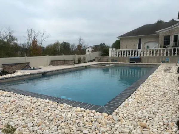 Backyard swimming pool with stone deck and house patio under an overcast sky