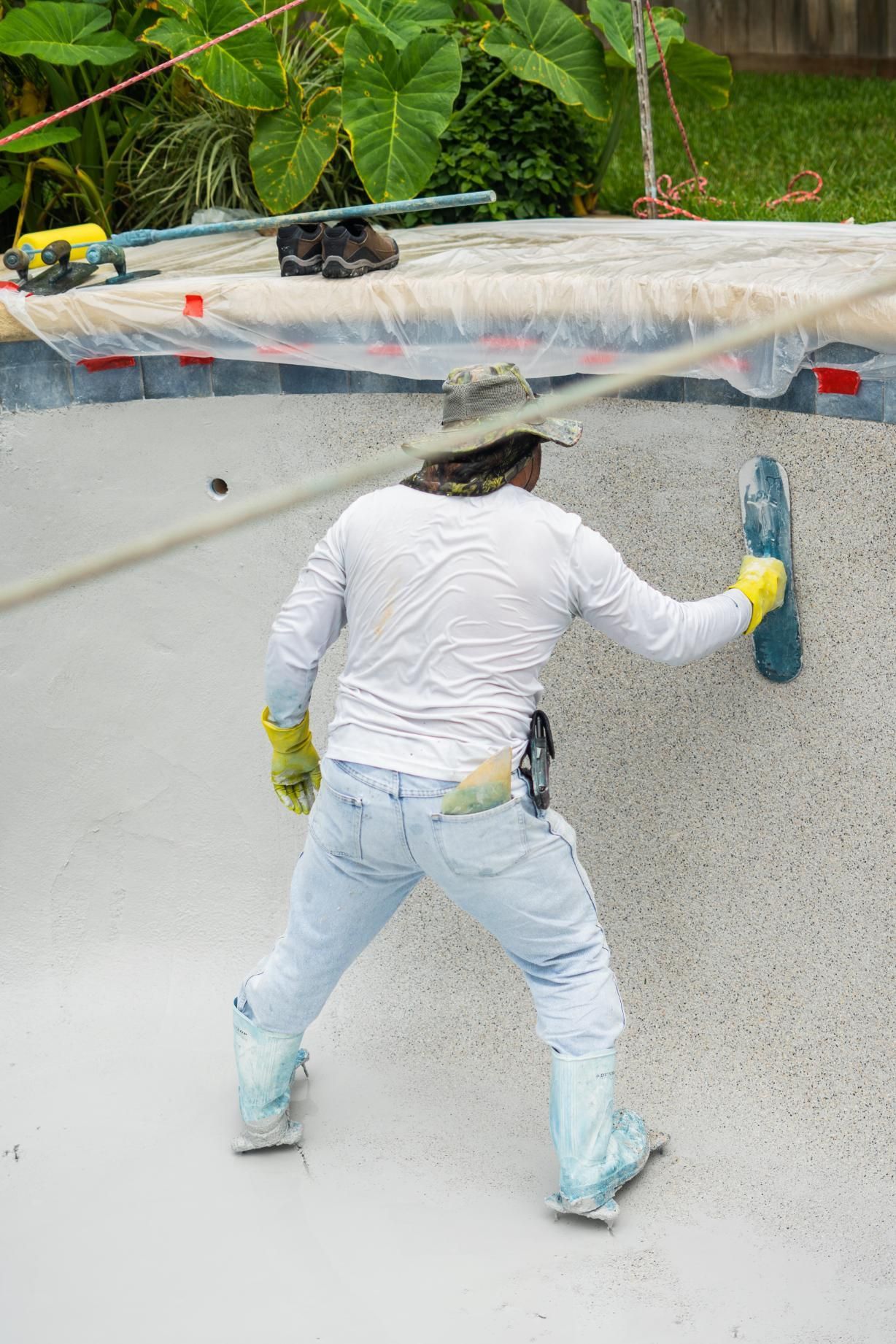 Worker smoothing white pool plaster with a trowel in a backyard pool under plastic sheeting