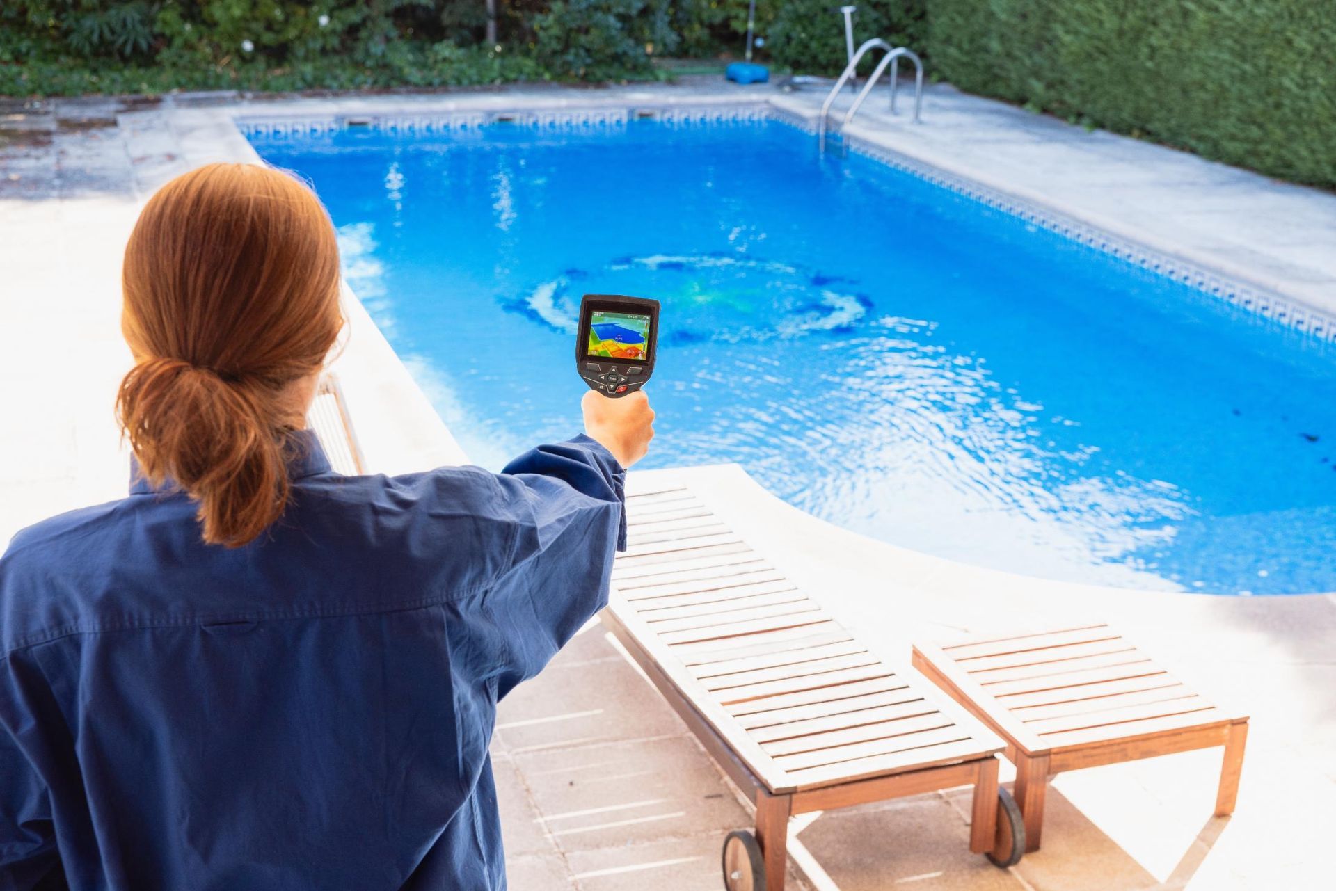 Person measuring pool water with a handheld tester beside a blue swimming pool and lounge chair