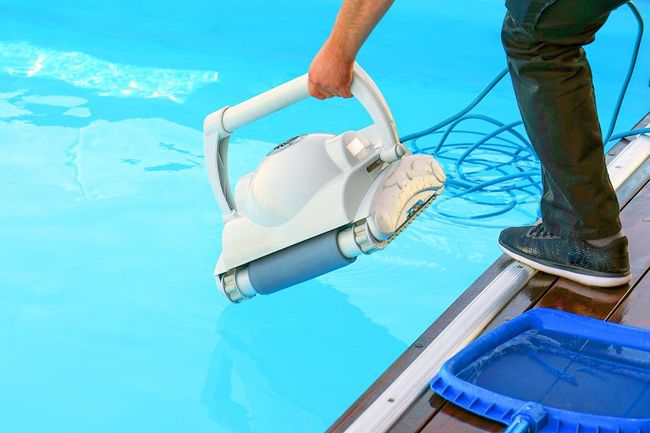 Person lowering a white pool vacuum into a bright blue swimming pool beside the deck