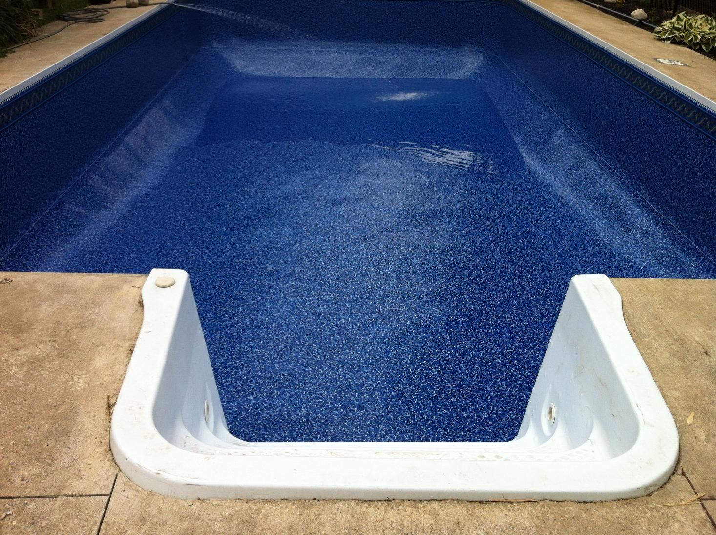 Empty backyard swimming pool with blue water, white steps, and tan stone deck