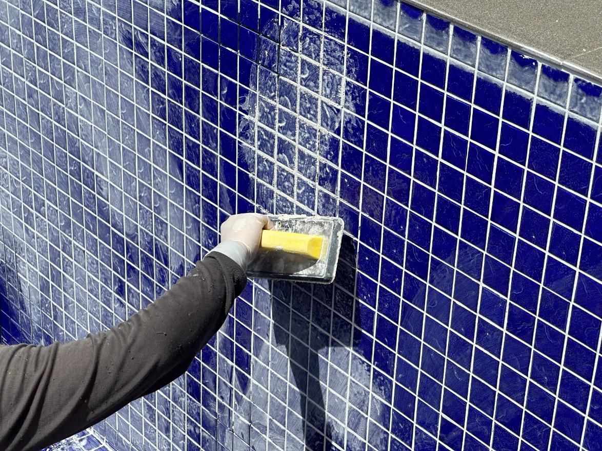 Hand trowel smoothing wet mortar on a blue-tiled wall beneath a metal grid