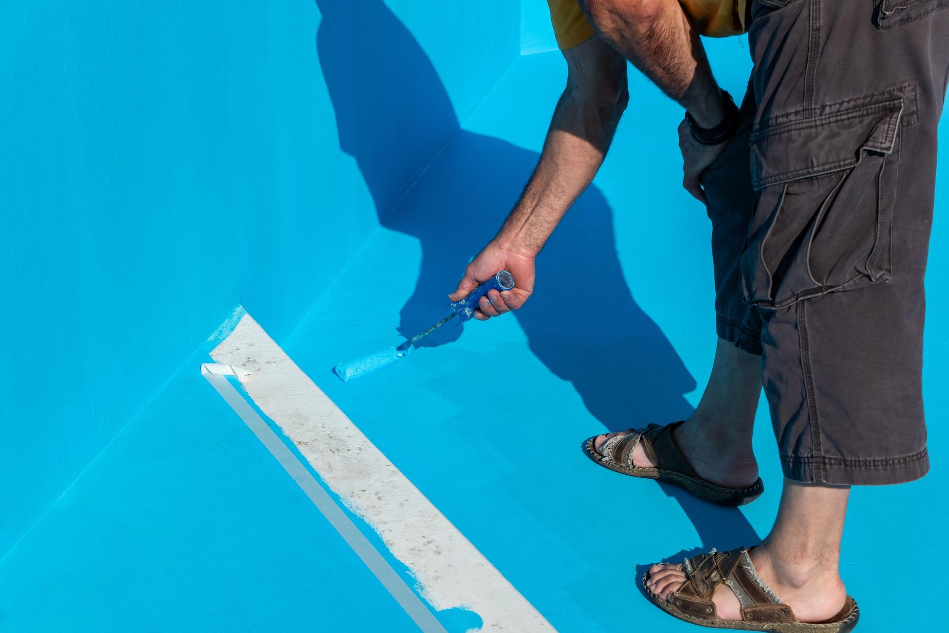 Person painting a bright blue surface with a roller, wearing dark shorts and sandals