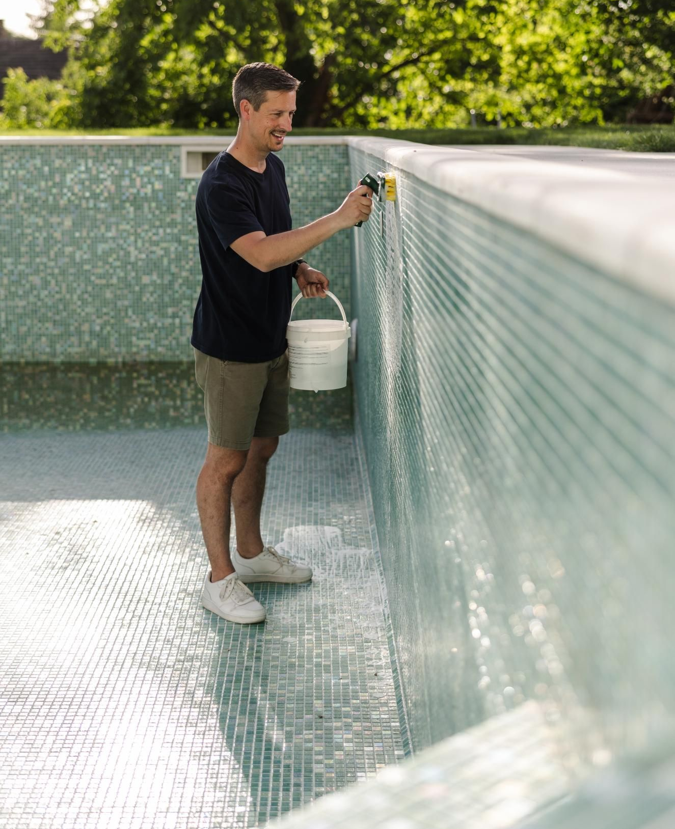 Person filling a white bucket at the edge of an empty pool with blue mosaic tiles