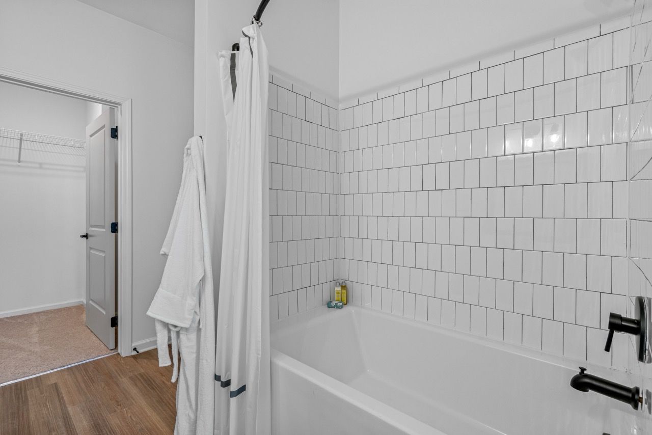 Bathroom with white tiled shower surround, bathtub, shower curtain, and robe hanging by the door.