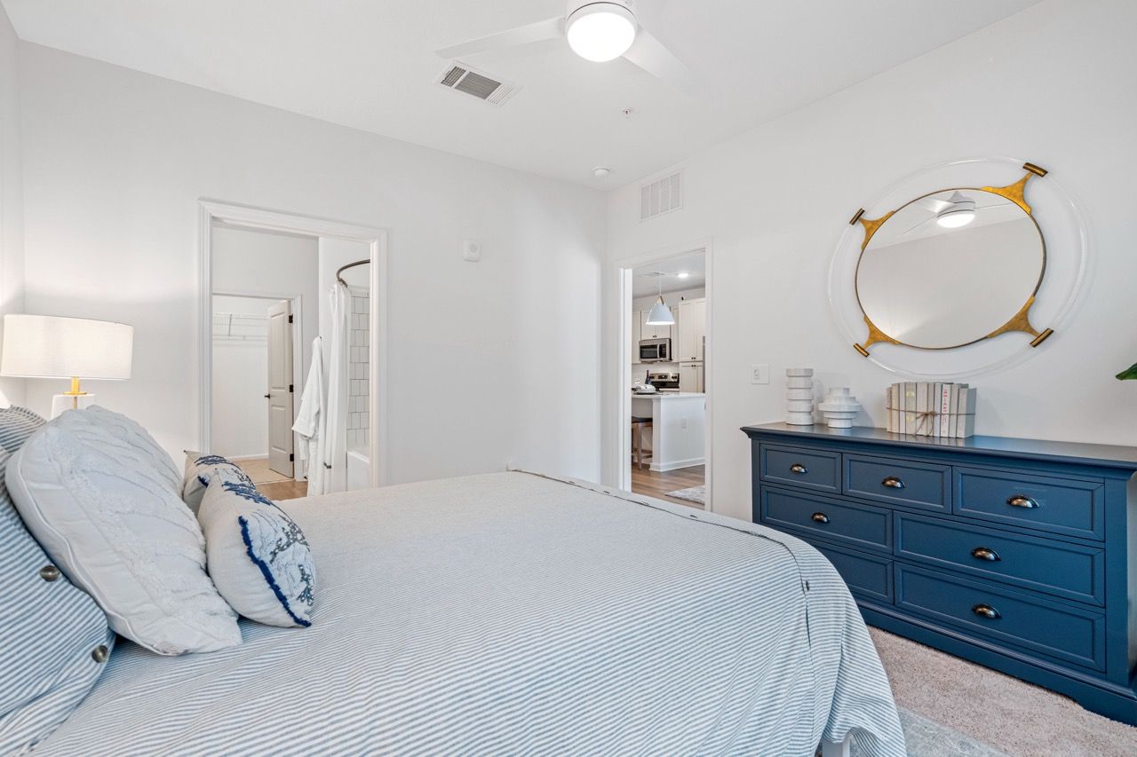 Bright bedroom with striped bedding, blue dresser, round mirror, and doorway to the kitchen.