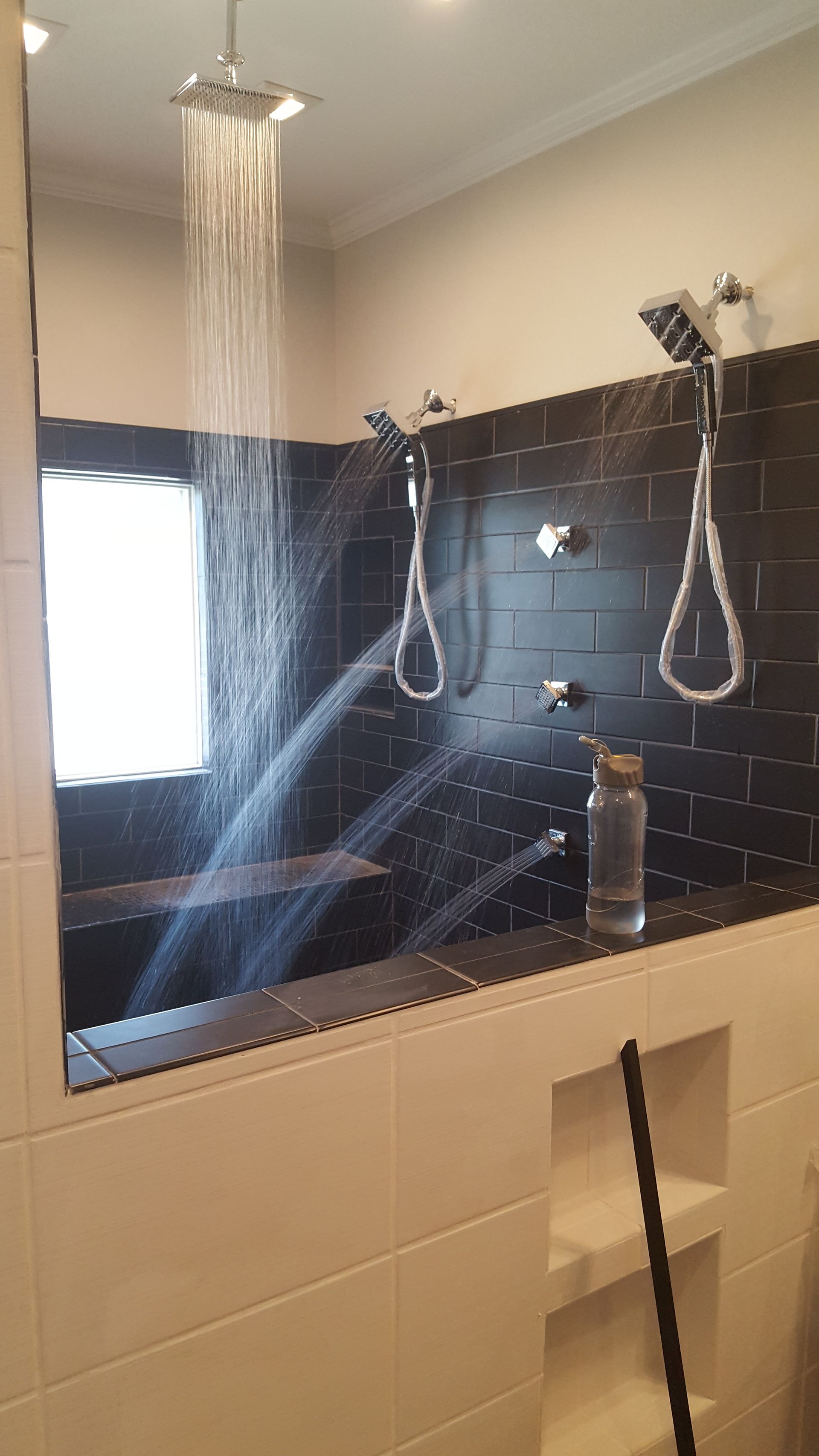 Bathroom shower with dark tile, rainfall showerhead, two hanging showerheads, and sunlight through a window