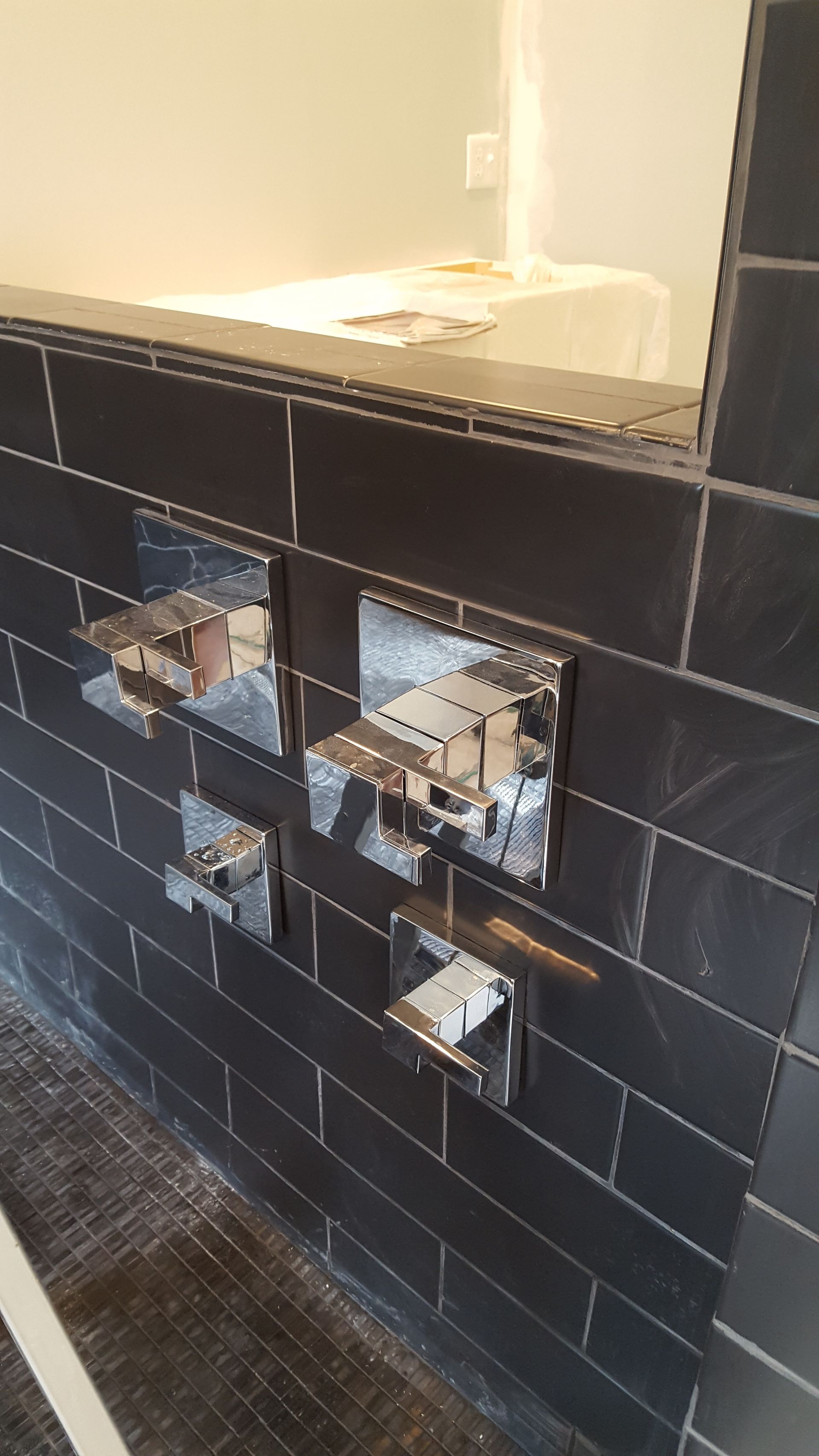 Black-tiled bathroom wall with multiple exposed chrome plumbing fixtures and sink openings