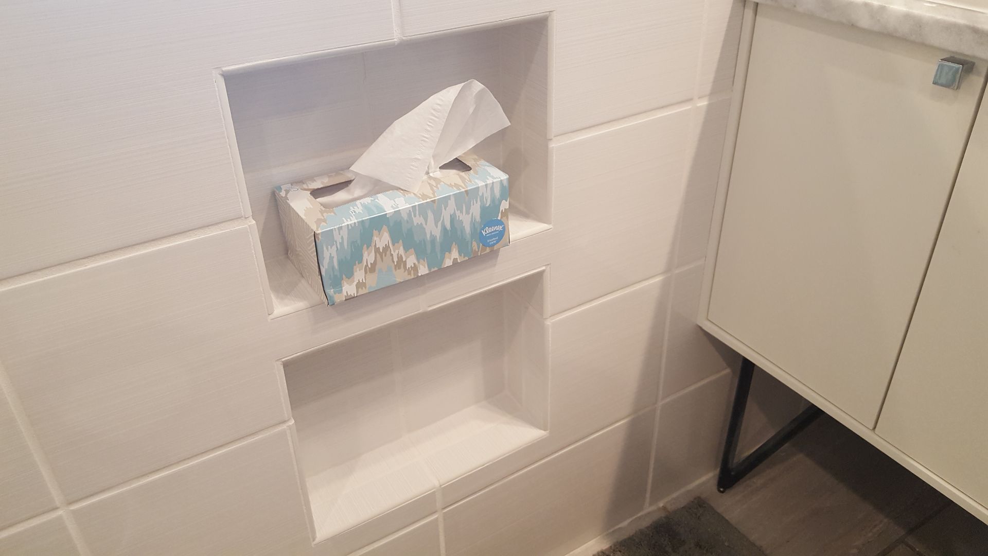 Wall-mounted tissue box in a white tiled bathroom beside a cabinet.