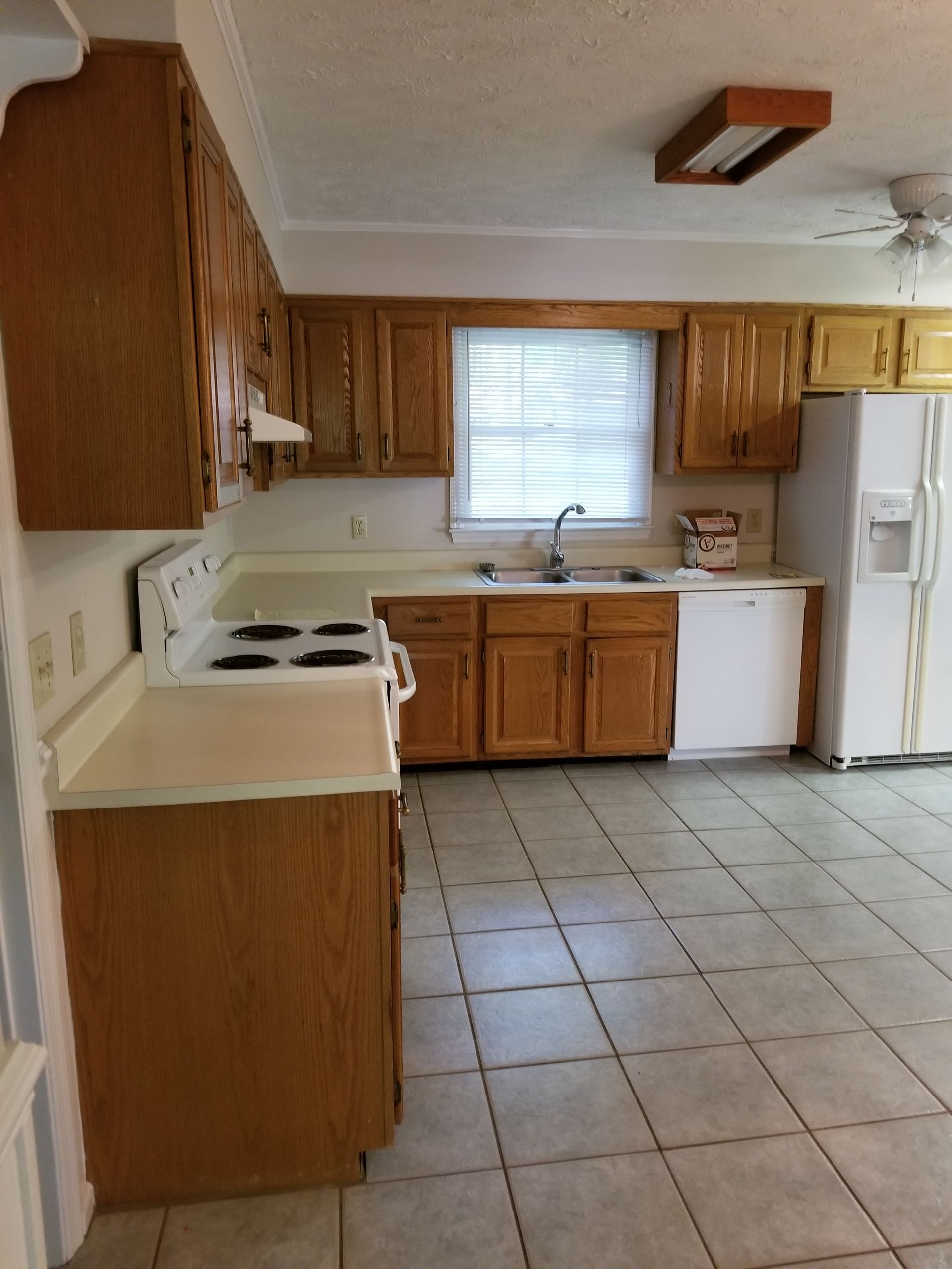 Empty kitchen with wood cabinets, white appliances, tiled floor, and a window over the sink.